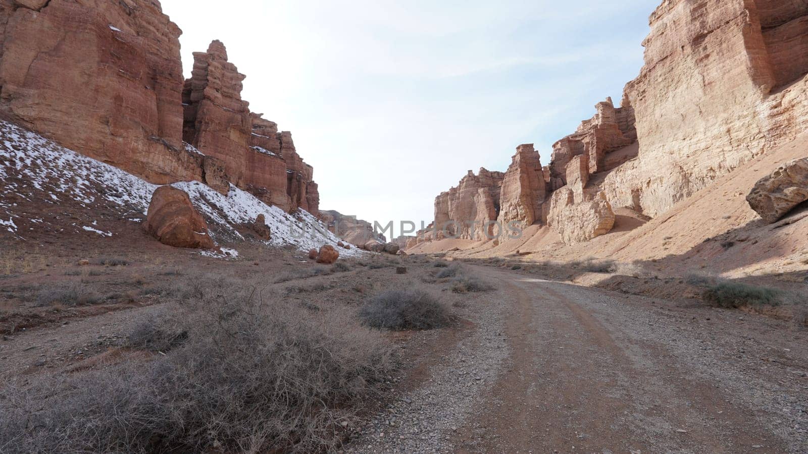 Grand Canyon Charyn. Orange cliffs and rocks. In places there are dry bushes and there is snow. The sun is shining brightly. White clouds. Faults and cracks in the canyon. The place looks like Mars