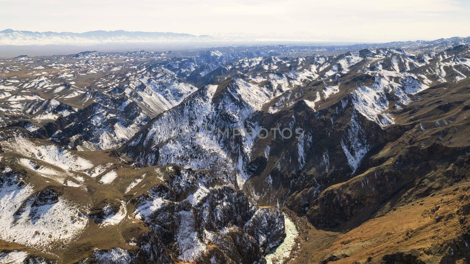 A long river with green water in the Charyn Canyon. There is white snow in places. Bushes and trees grow. The river runs through a canyon among rocks and cliffs. Mountains are visible. Drone view