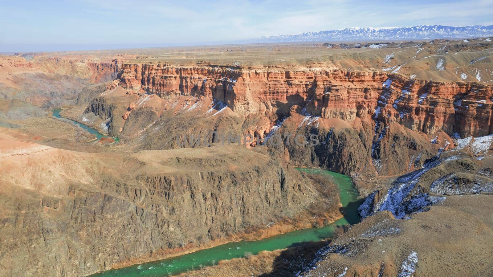 A long river with green water in the Charyn Canyon. There is white snow in places. Bushes and trees grow. The river runs through a canyon among rocks and cliffs. Mountains are visible. Drone view
