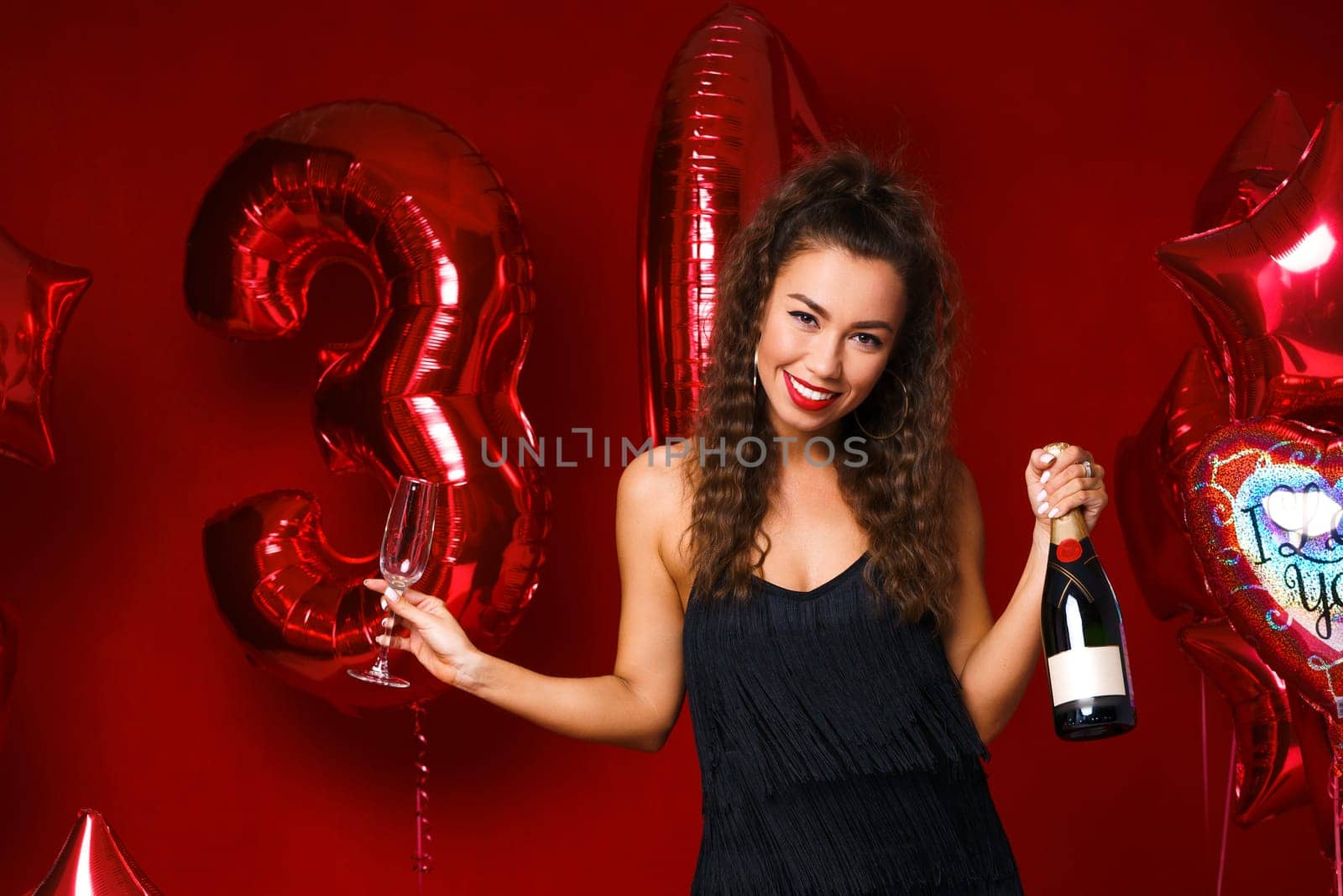 Happy woman with bottle of champagne on red background with red balloons. Cheerful mood at the holiday, anniversary of 30 years. A girl with long hair and bright red lips smiles with snow-white teeth