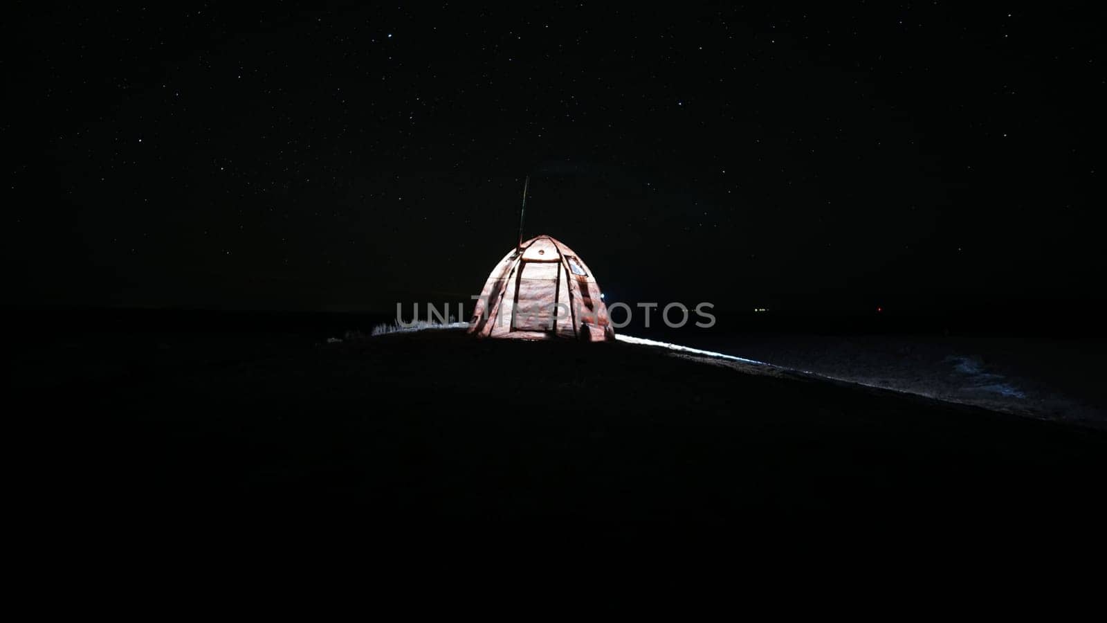 A tent with a stove stands under the starry sky. Night. Bright stars. Portable stove in the tent. The light is on. Lights are visible in the distance. Grass grows on the ground and snow lies in places