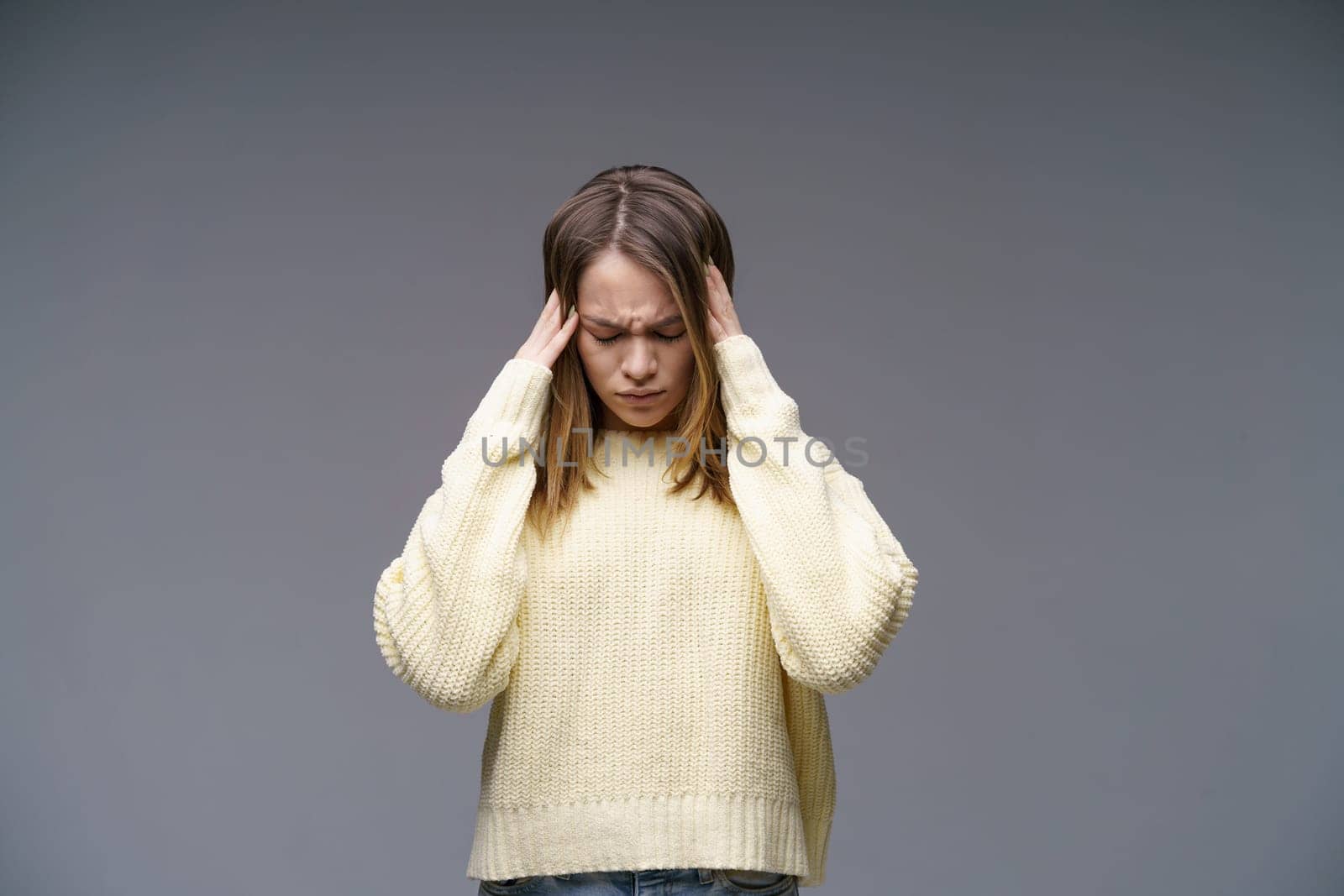 Young woman holding her head in yellow sweater on gray background by EkaterinaPereslavtseva