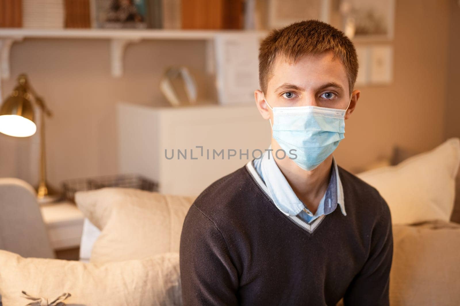 a young guy of Caucasian ethnicity in a shirt and a protective mask sits on the couch alone at home. Concept of being at home during global quarantine