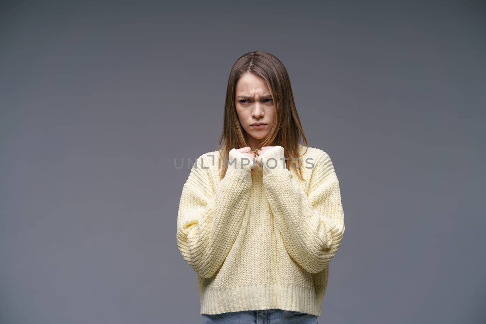 Young upset woman in yellow sweater on gray background holding hands in fists by EkaterinaPereslavtseva