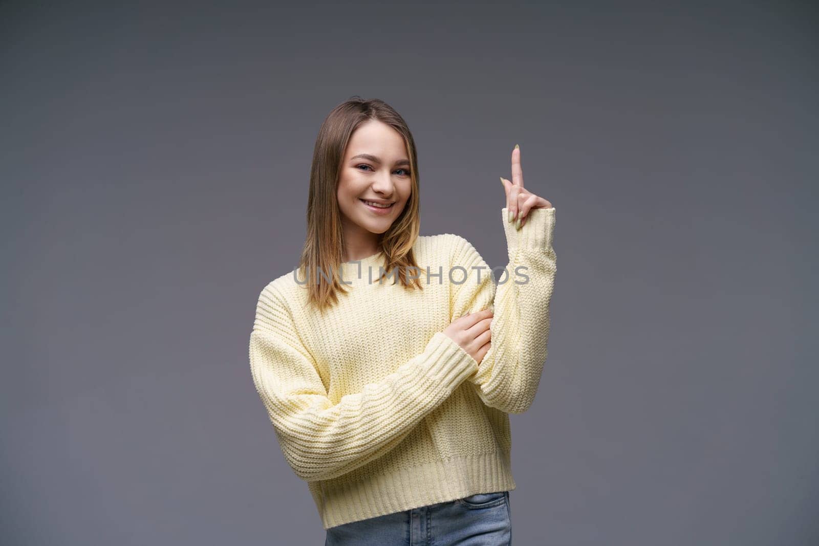 Cheerful young woman of Caucasian ethnicity shows thumbs up in a yellow sweater on a gray background, positive advertising. Beautiful girl posing in studio