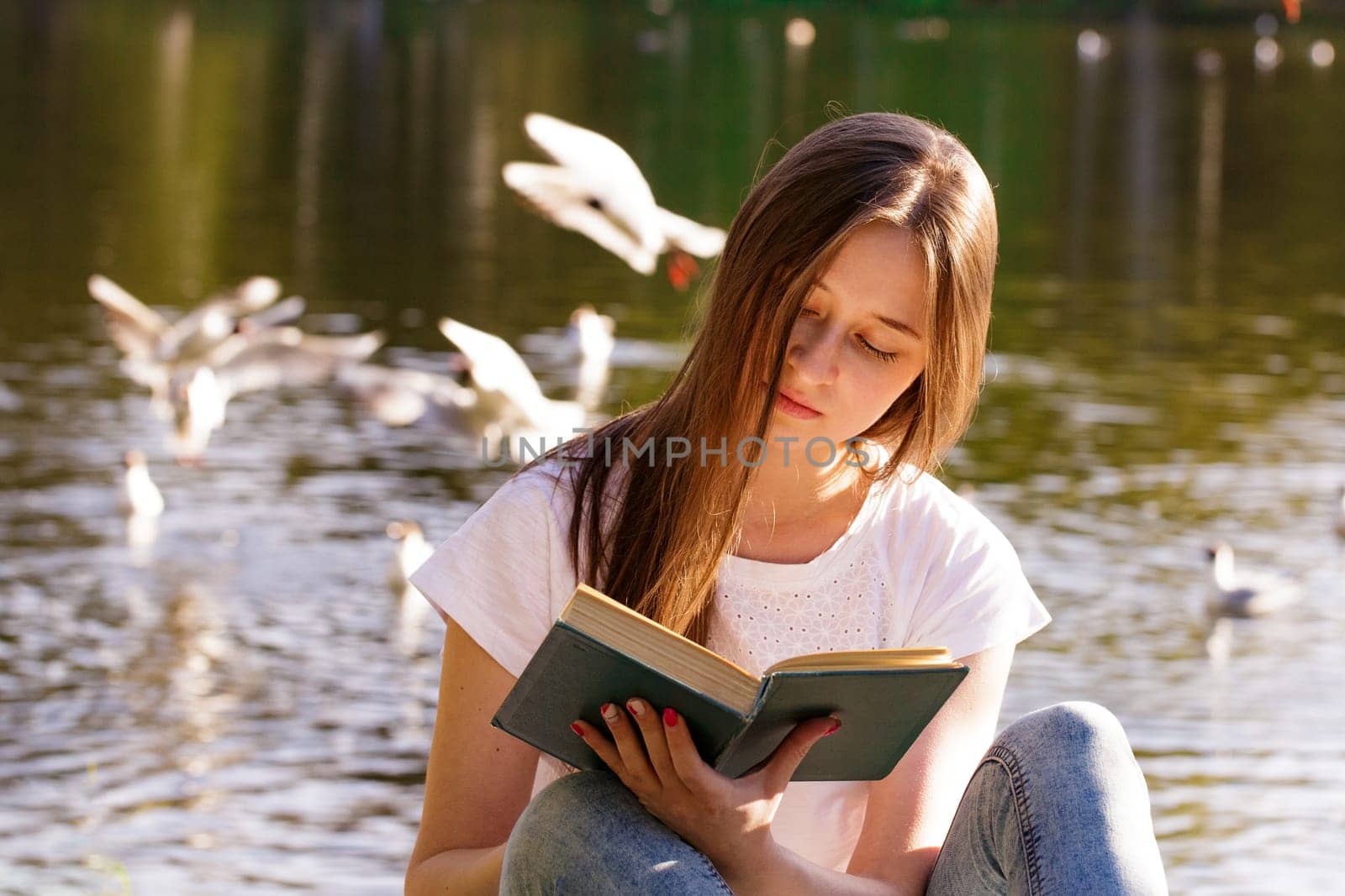 Young caucasian woman sitting on the shore of the lake reads a book on a sunny day. Reads outdoors in the park