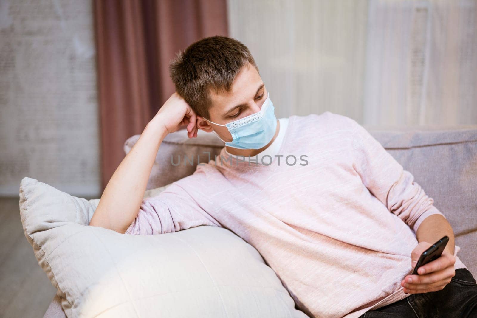 young caucasian man sitting on a sofa in casual clothes, wearing a protective medical mask with a phone in his hand