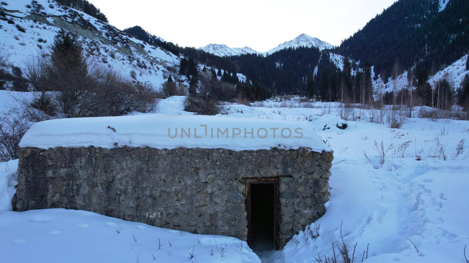 An old stone prison or hut in the mountains. Snowy mountains, forest and clouds against a blue sky. Tall dry bushes peek out from under the snow. Birch and spruce trees are visible in the distance.