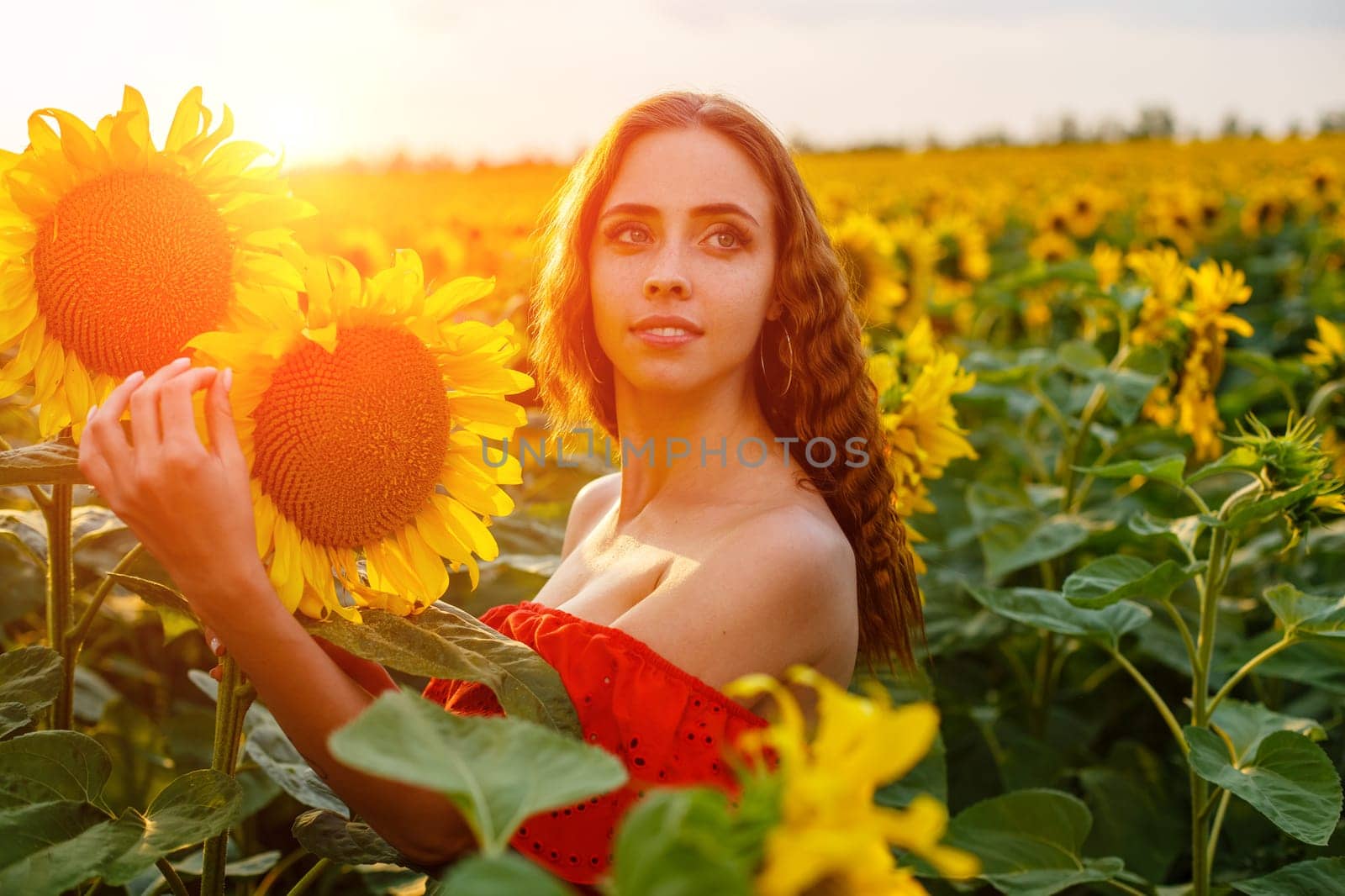 Beautiful curly young woman in sunflower field holding sunflower flower in hand . Portrait of young woman in sun smiling cute. Charming girl of Caucasian appearance with wavy hair in a red dress