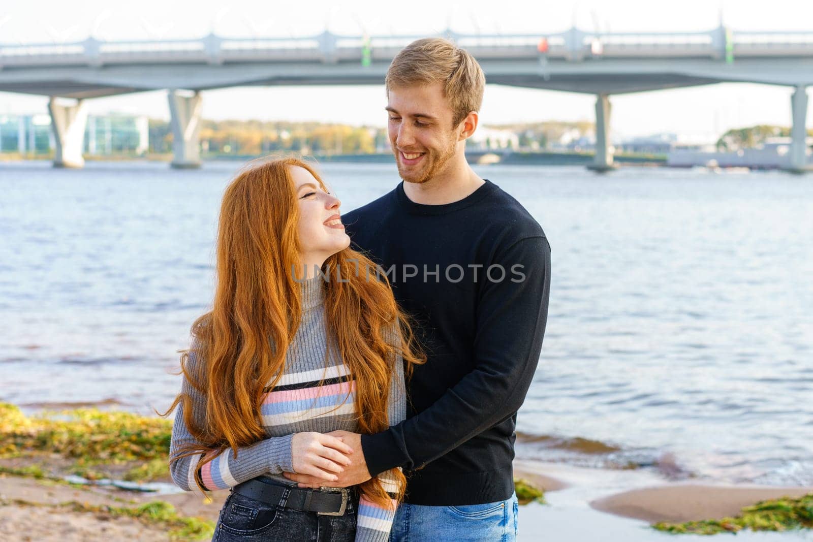 Young couple of man and woman with long red hair of Caucasian ethnicity, in casual clothes, stand on the bank of the river embracing happy on a summer day against the background of the cityscape