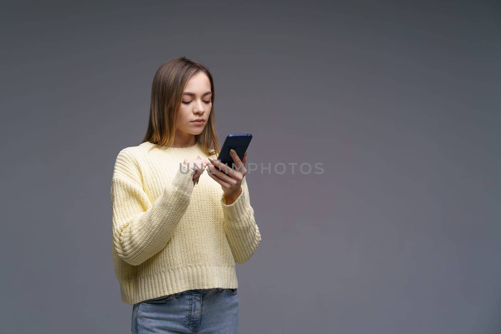 a young beautiful woman of Caucasian ethnicity writes sms on the phone, stands in a yellow sweater on a gray background, communicating via the Internet