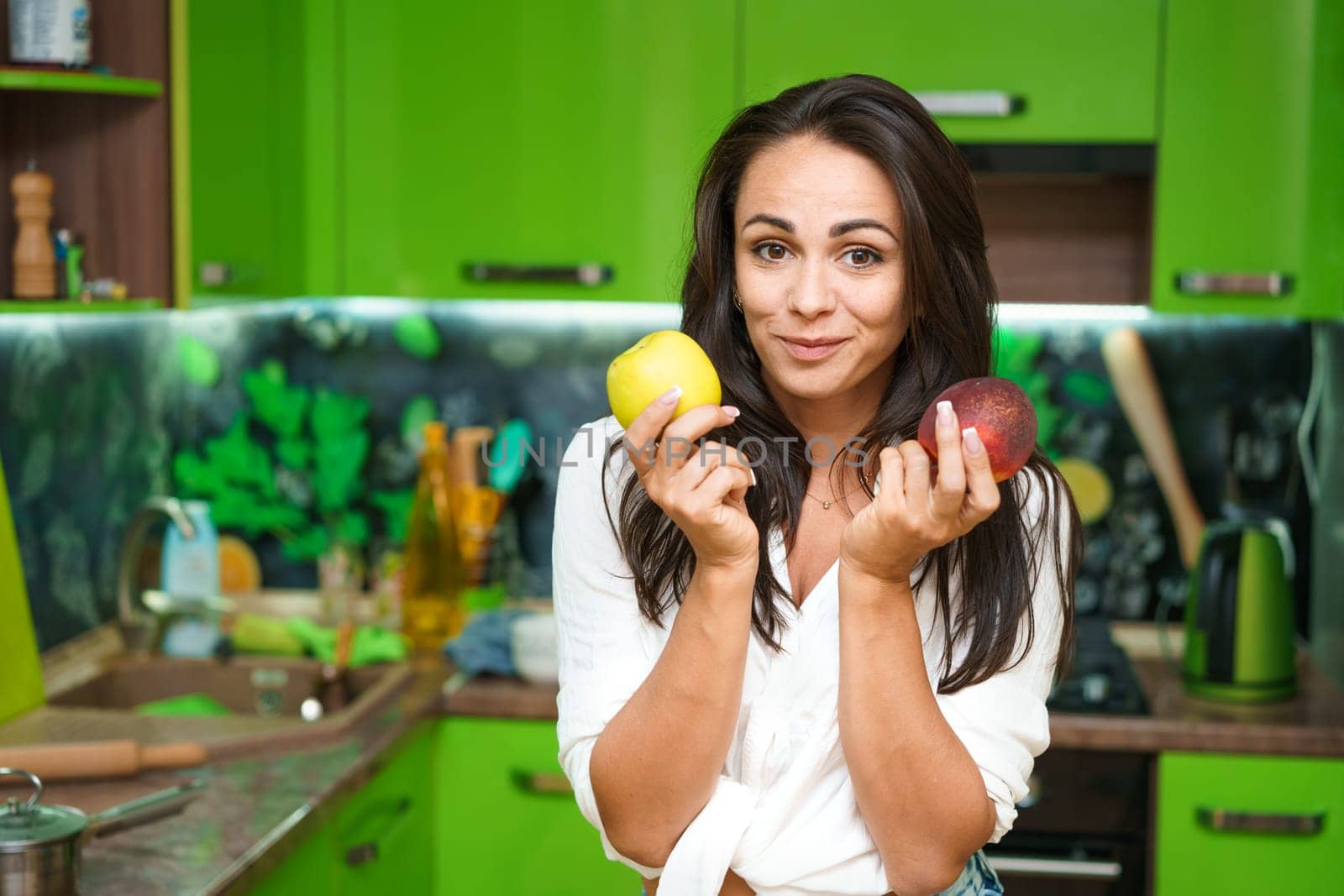 Young woman stands in kitchen holding fruits in her hands. Caucasian girl in a modern kitchen holds green apple and ripe peach in her hands. Chooses what to eat. Healthy nutrition for beautiful skin