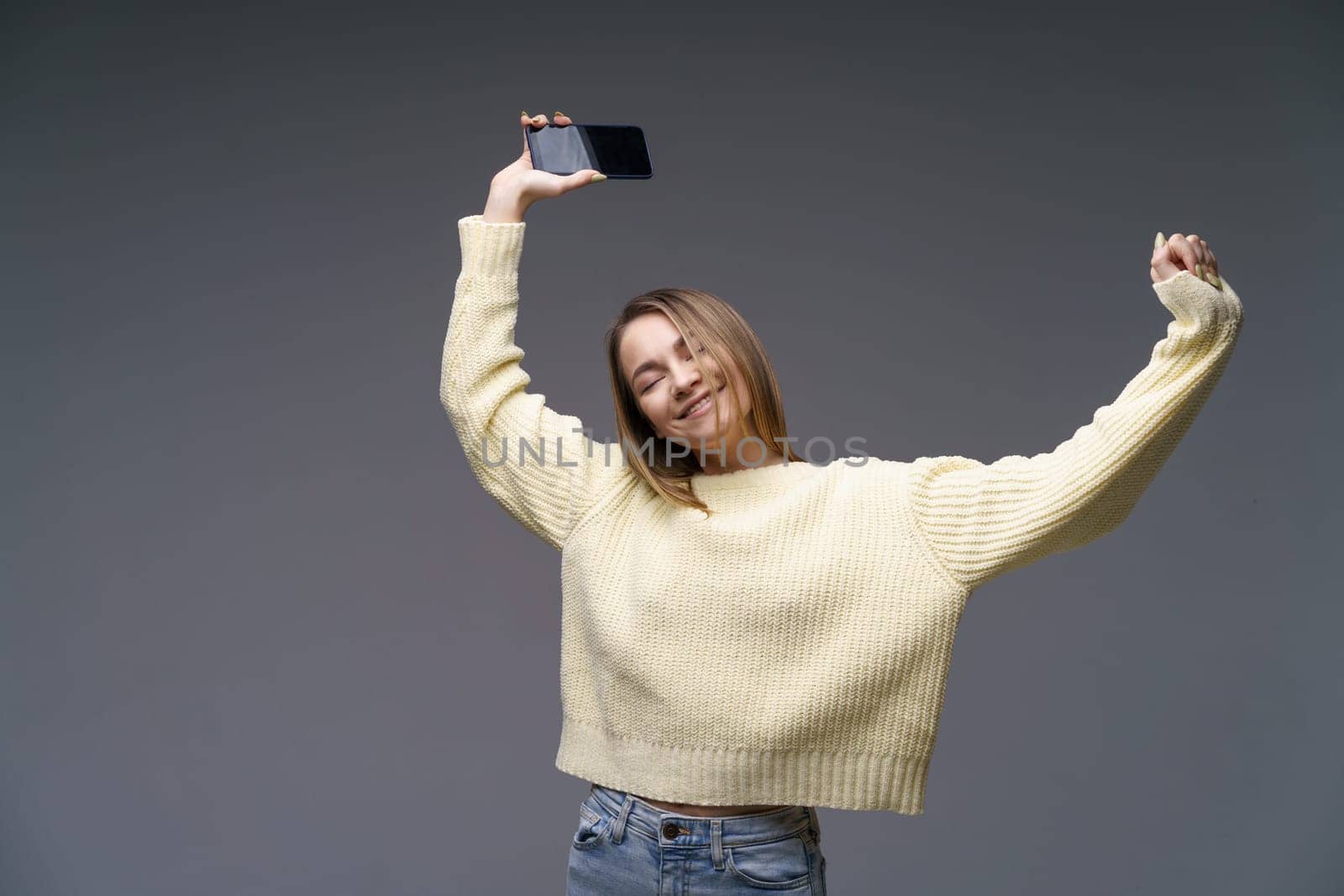 Young cheerful woman of Caucasian ethnicity in a yellow sweater on a gray background is dancing with a phone in her hand. Happy girl listening to music on the phone dancing raising her hands up