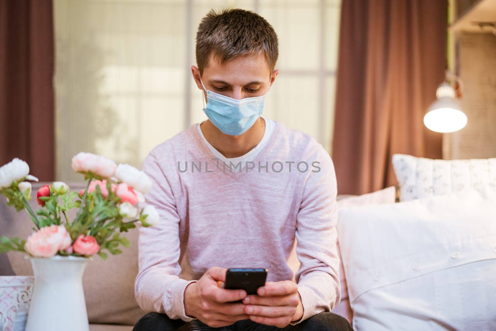 a young man sits on a sofa wearing a protective mask with a phone in his hand by EkaterinaPereslavtseva