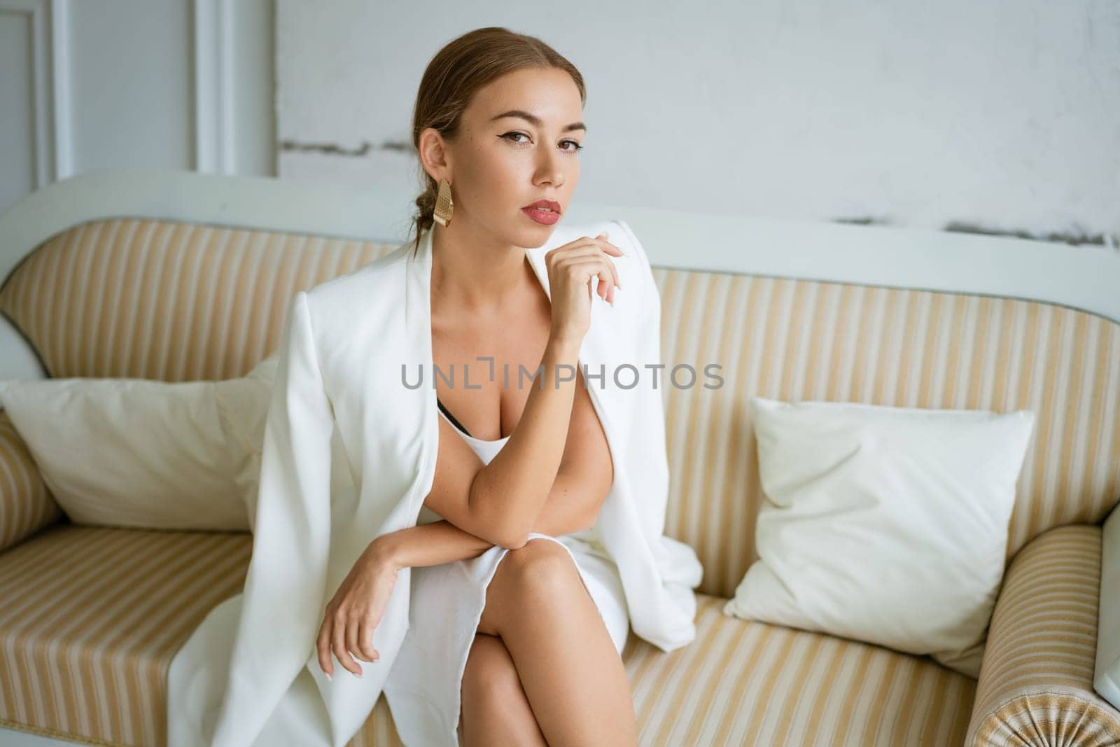Young business woman of Caucasian ethnicity is sitting on the sofa in white business clothes with beautiful makeup and hair. Portrait of a confident beautiful girl
