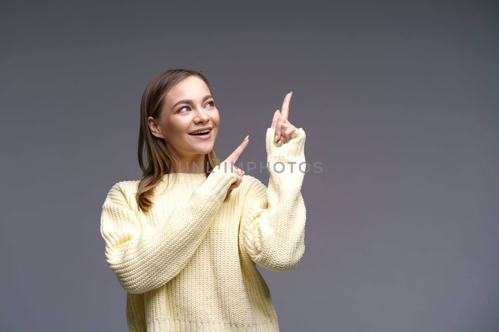 Cheerful young woman showing thumbs up in yellow sweater on gray background by EkaterinaPereslavtseva