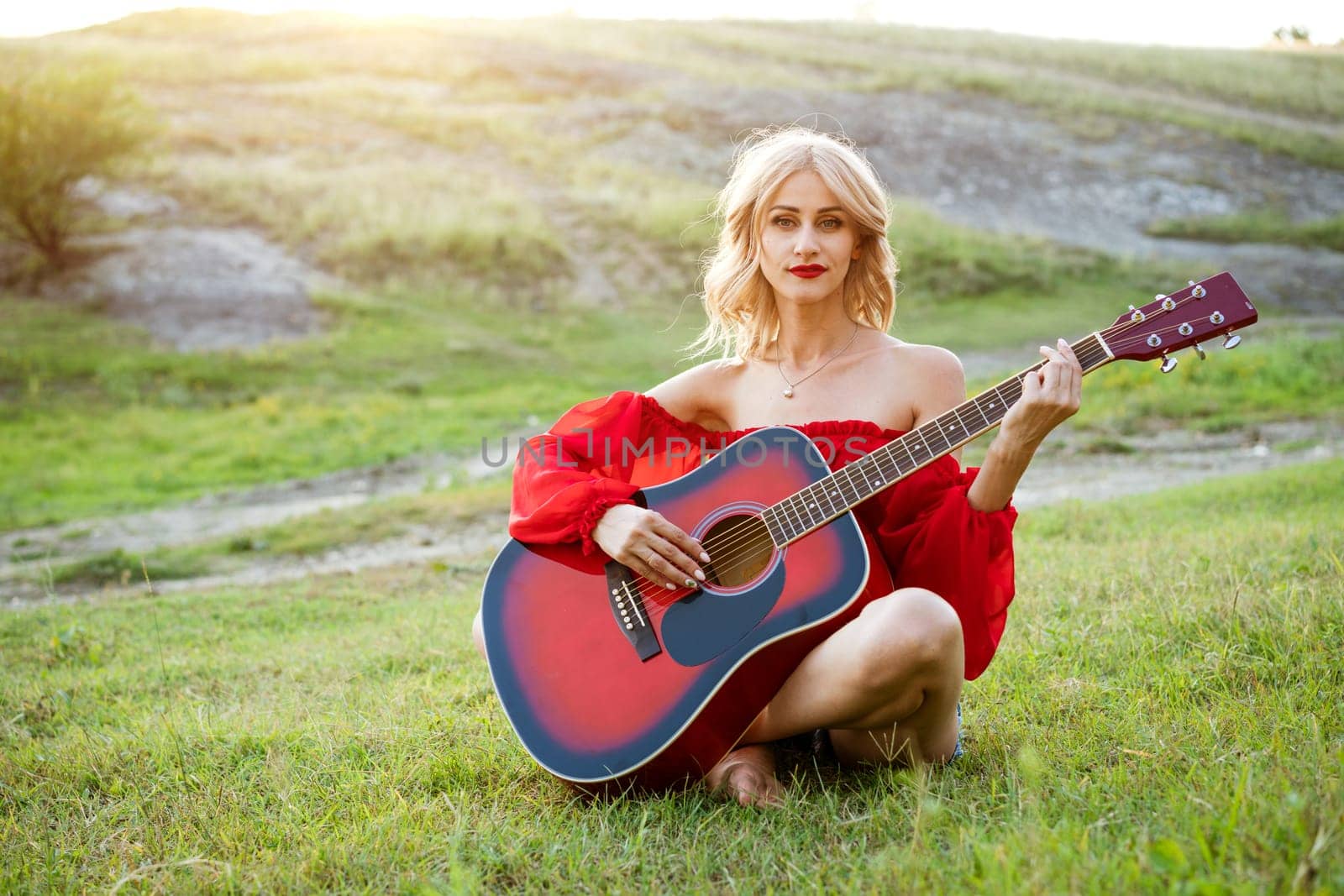 Woman in red grass with a red guitar. Girl musician plays the guitar outdoors. Blonde caucasian appearance