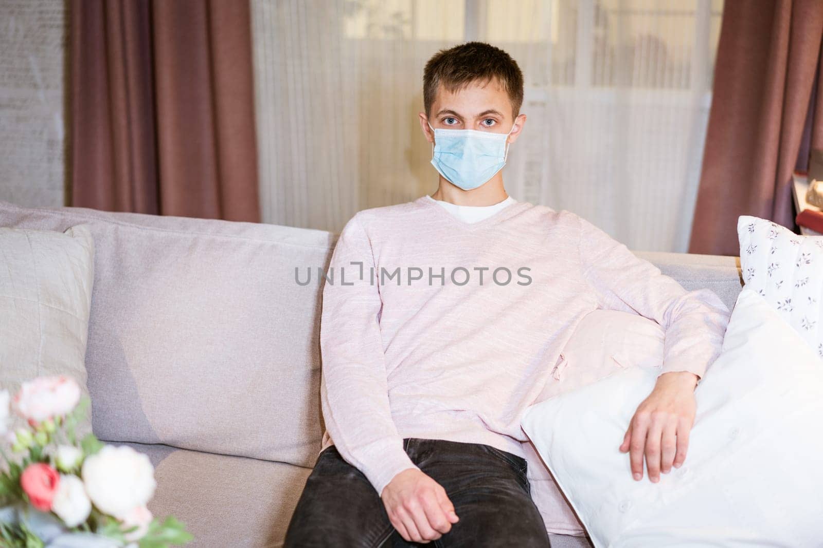 a young guy of Caucasian ethnicity in a shirt and a protective mask sits on the couch alone at home. Concept of being at home during global quarantine