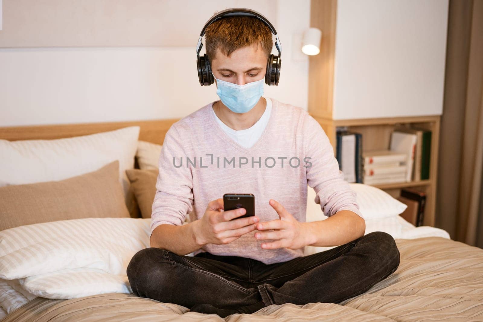 man of European appearance in casual clothes, wearing headphones and protective mask with phone his hands on bed at home, spending time Internet while sitting in quarantine during Lokdan