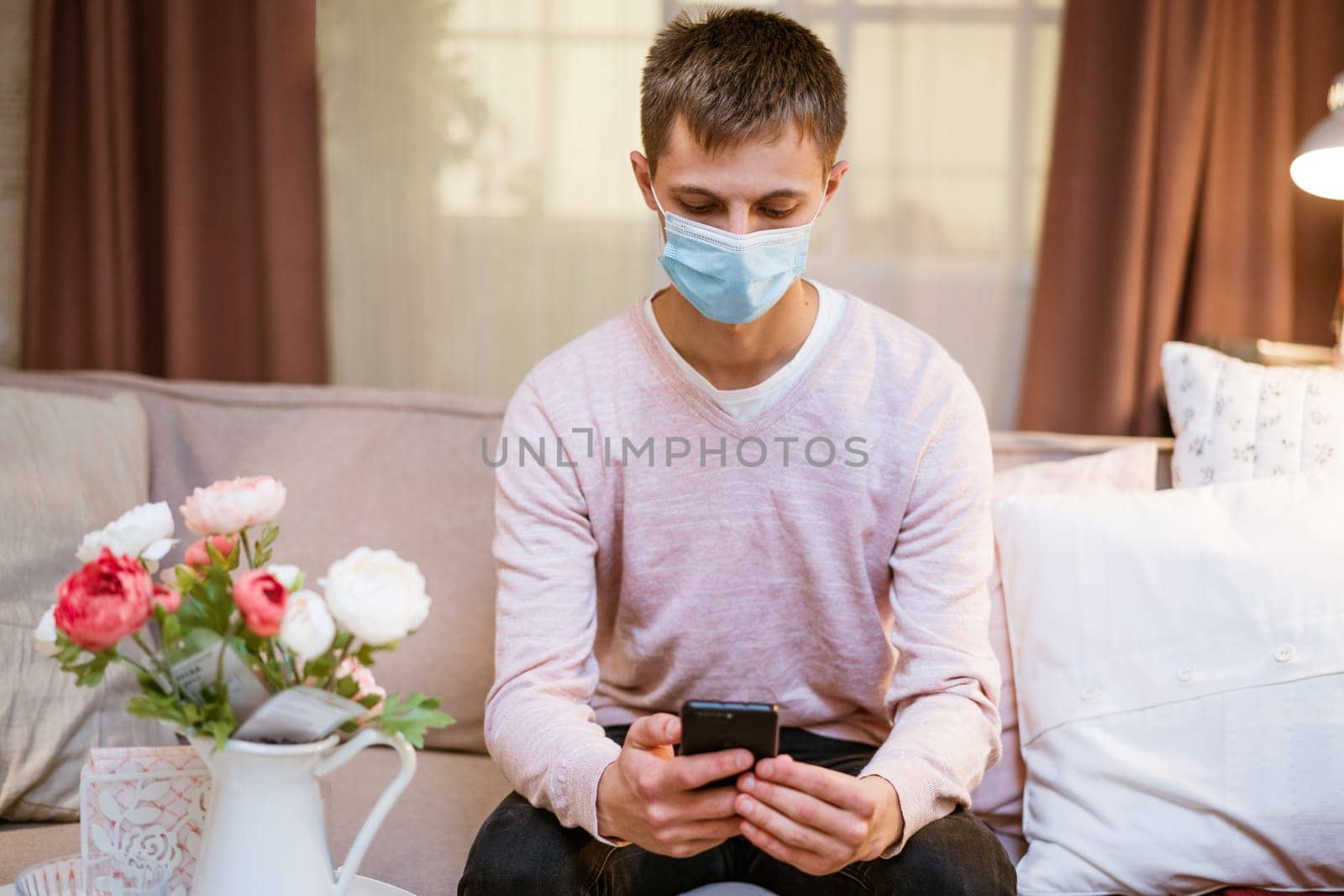 young man of European appearance in casual clothes, at home on the couch in a medical mask communicates by video communication on the phone, the concept of remote online communication on the Internet