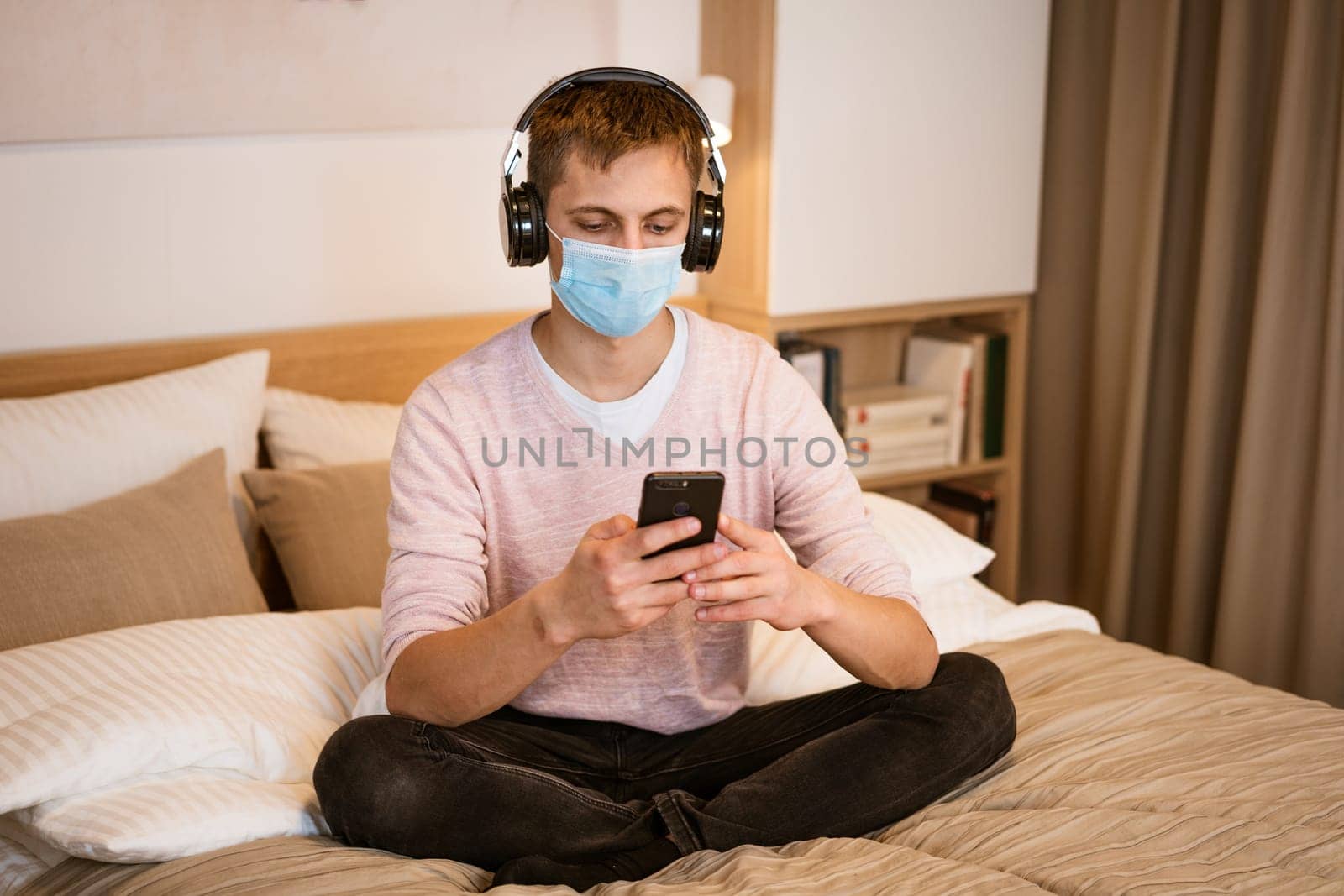 a guy of Caucasian nationality in casual clothes, wearing headphones on the bed in a protective medical mask with a phone in his hand, watching a video online on a Wi-Fi network