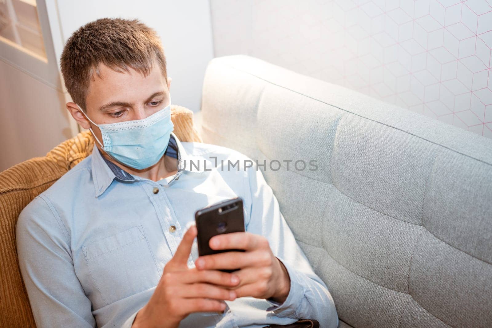 young caucasian man sitting on a sofa in casual clothes, wearing a protective medical mask with a phone in his hand