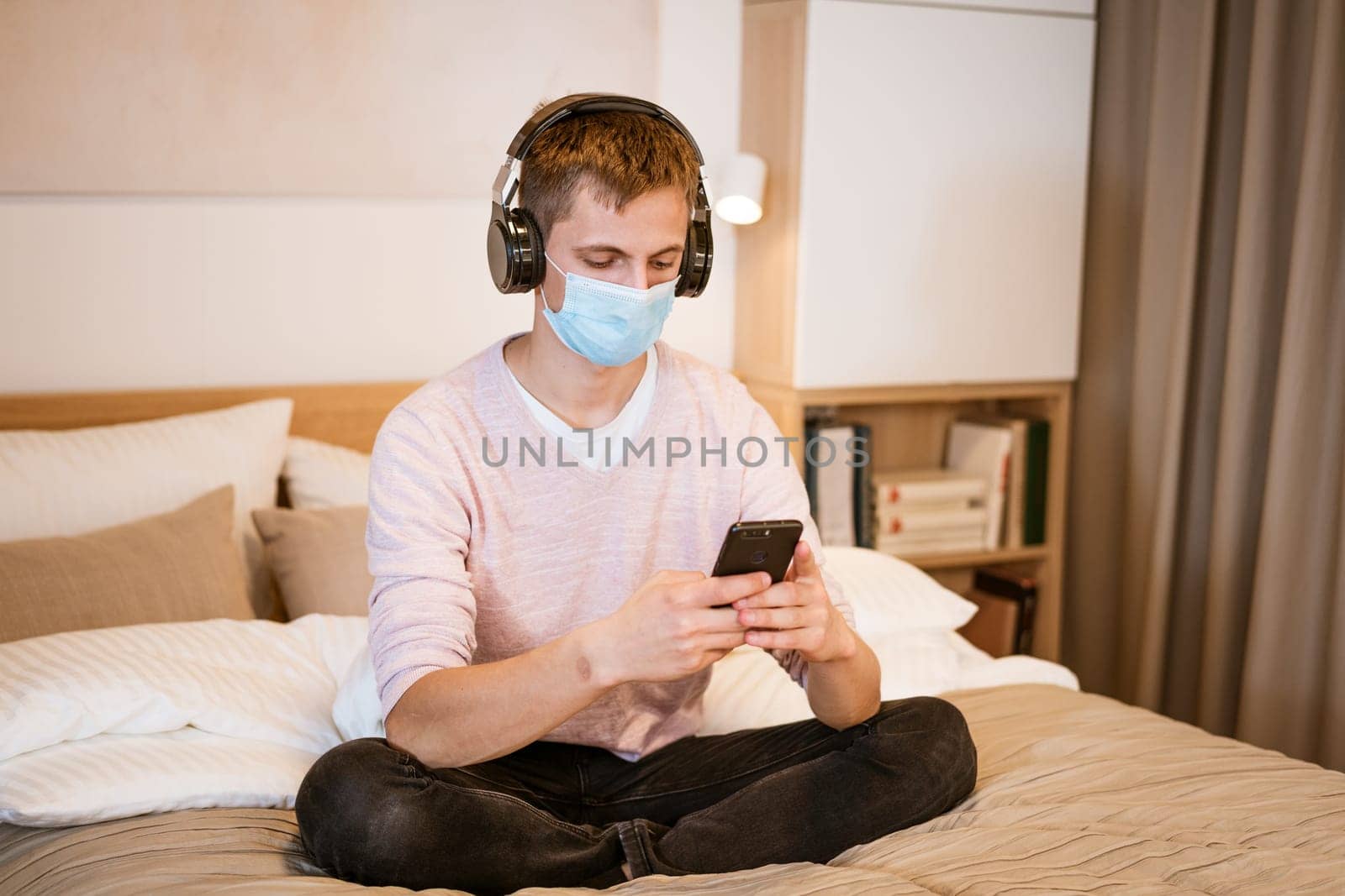 a guy of Caucasian nationality in casual clothes, wearing headphones on the bed in a protective medical mask with a phone in his hand, watching a video online on a Wi-Fi network