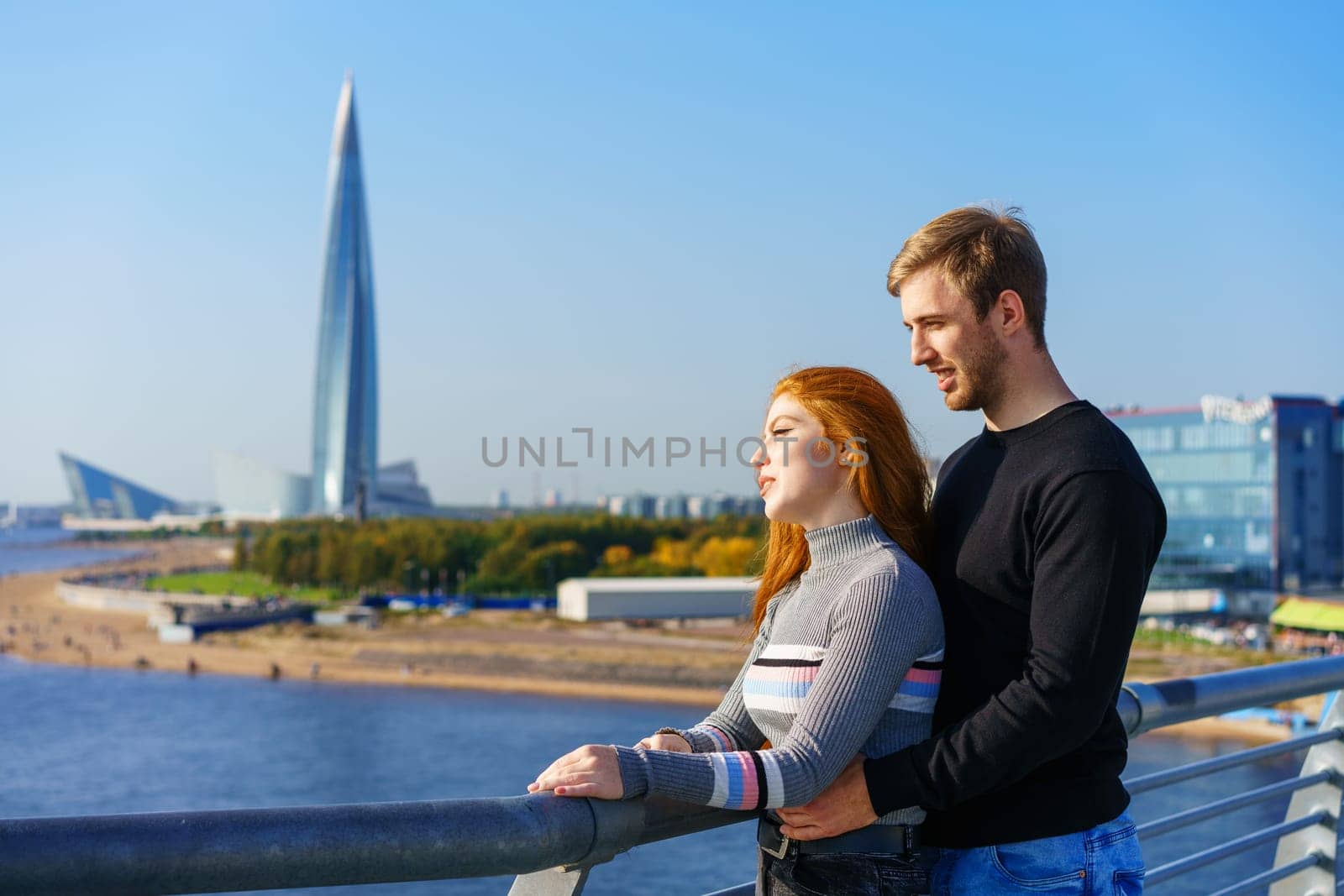 A guy and a girl of Caucasian ethnicity stand together on a river bridge on a sunny day with a city view