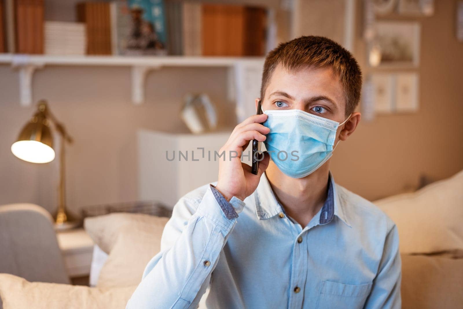 a young man of Caucasian appearance, in casual clothes, talking on the phone in a protective mask while sitting on the couch at home. The concept of remote communication