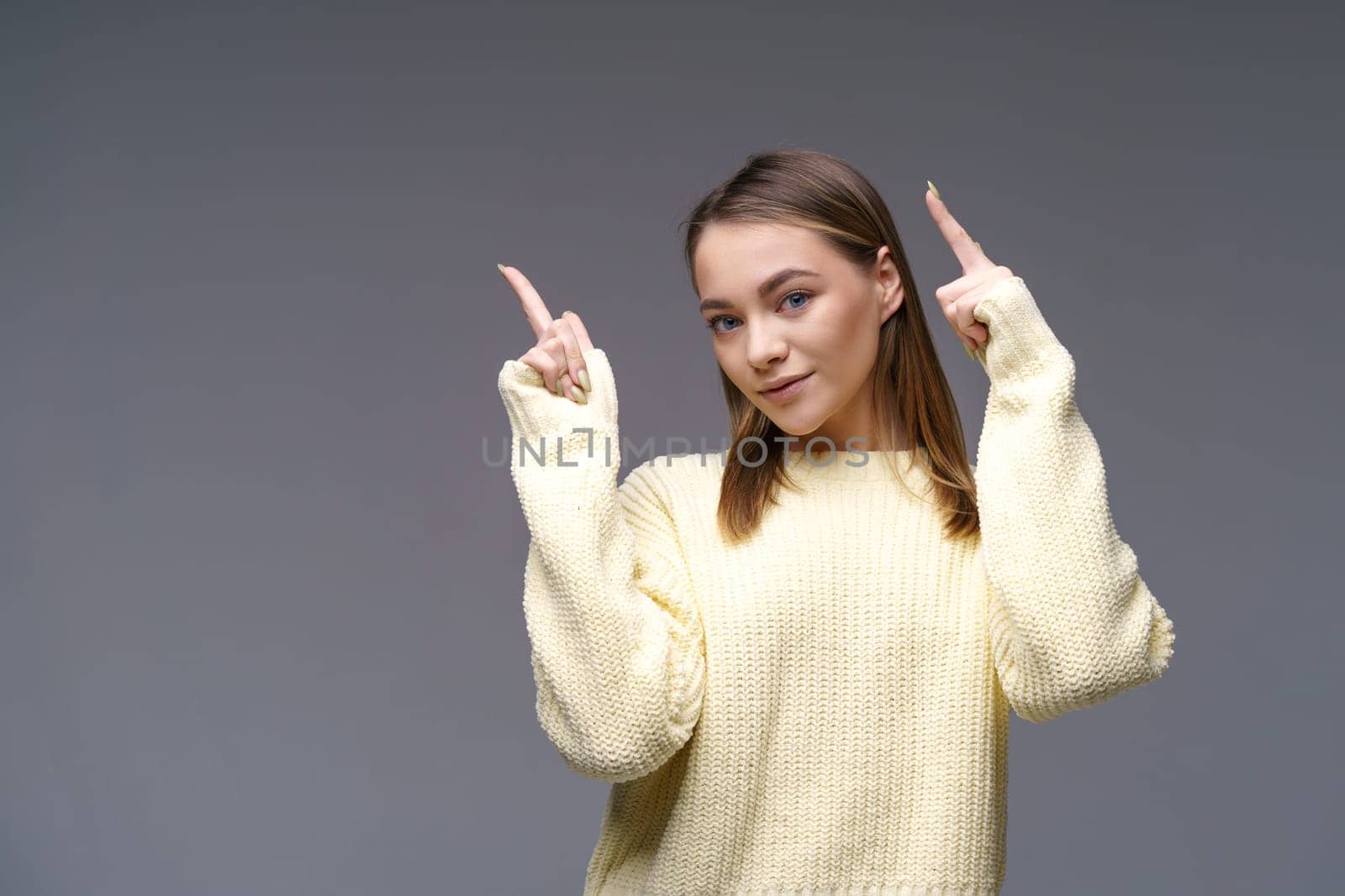 Cheerful young woman of Caucasian ethnicity shows thumbs up in a yellow sweater on a gray background, positive advertising. Beautiful girl posing in studio