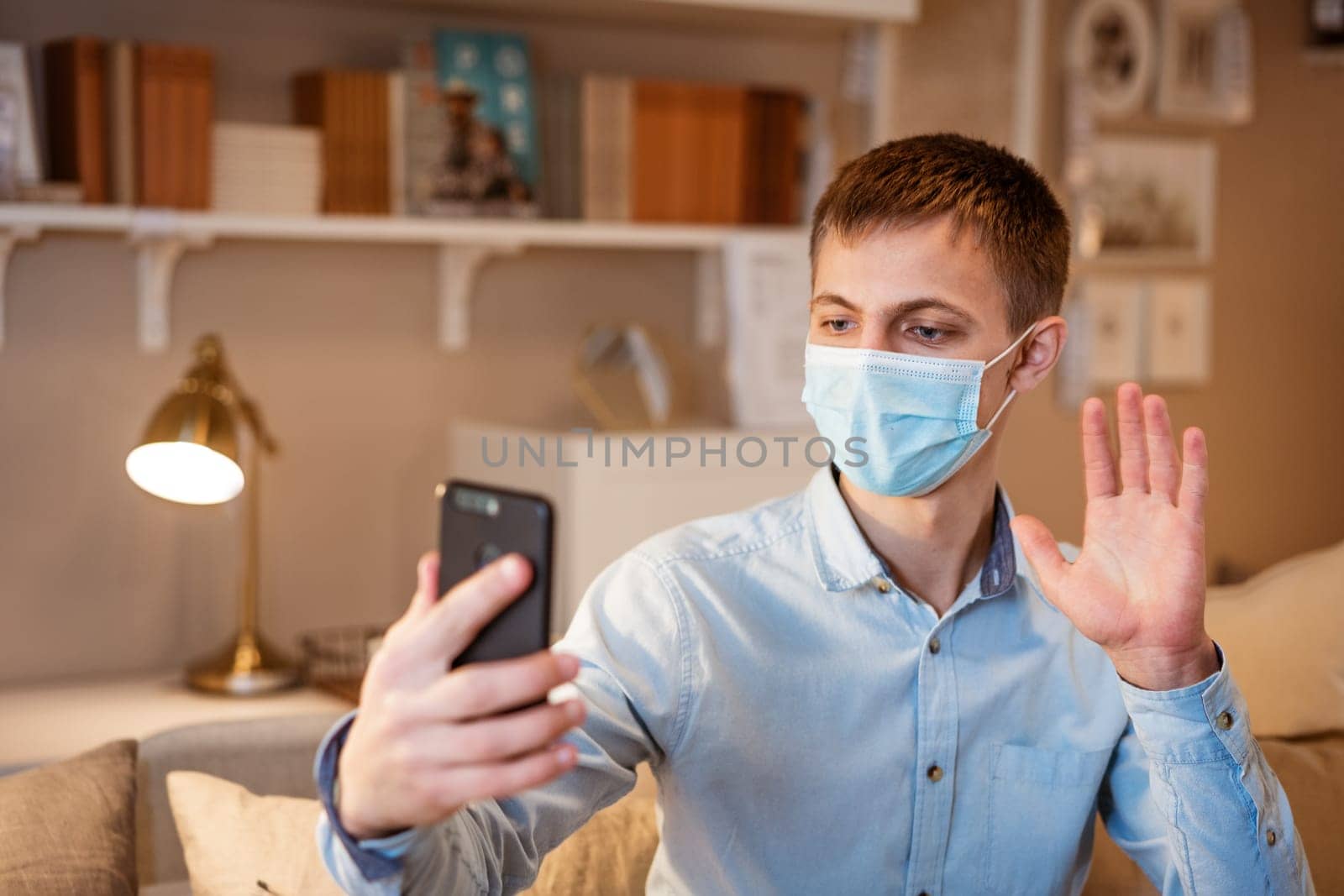 young man of European appearance in casual clothes, at home on the couch in a medical mask communicates by video communication on the phone, the concept of remote online communication on the Internet