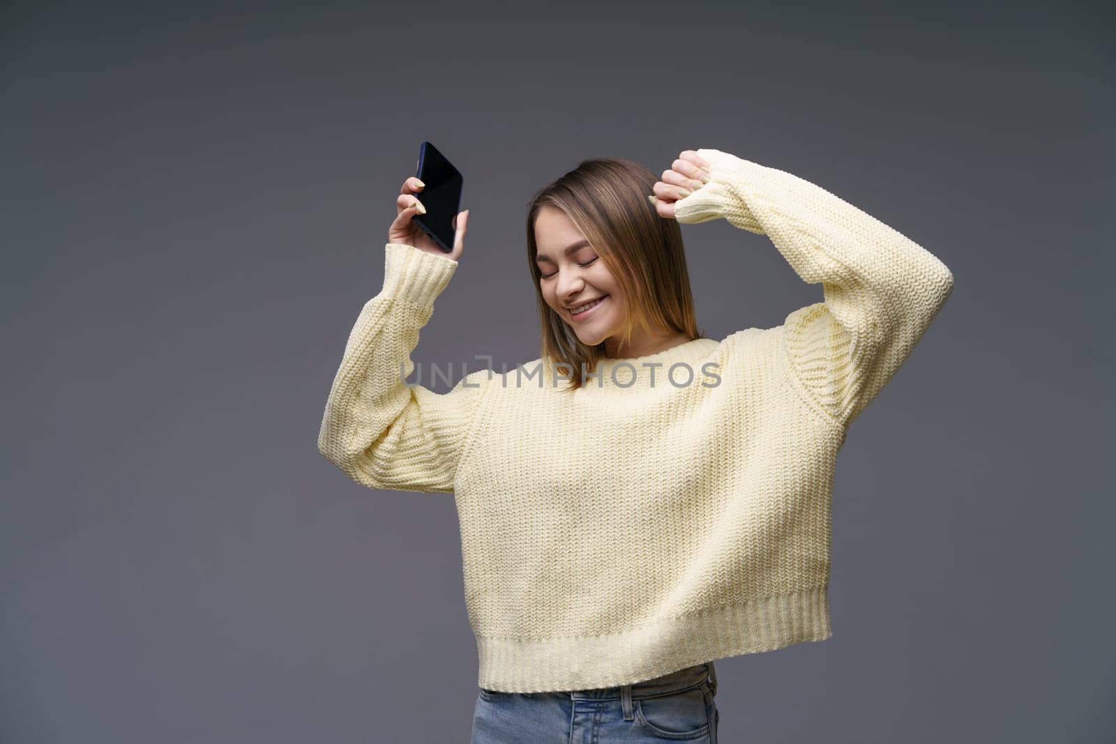 Young woman in yellow sweater on gray background dancing with phone in her hand by EkaterinaPereslavtseva