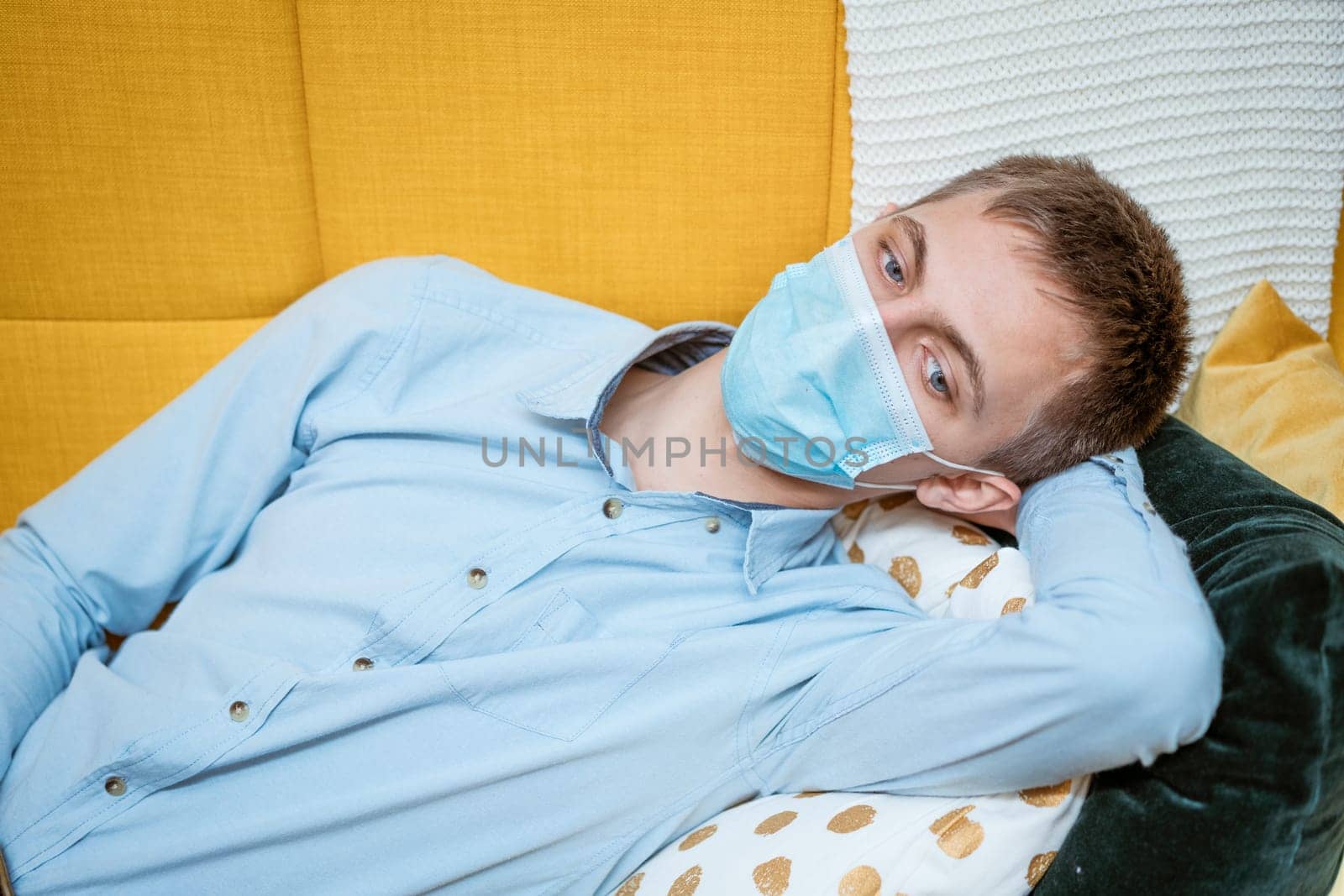 a young guy of Caucasian ethnicity in a shirt and a protective mask sits on the couch alone at home. Concept of being at home during global quarantine