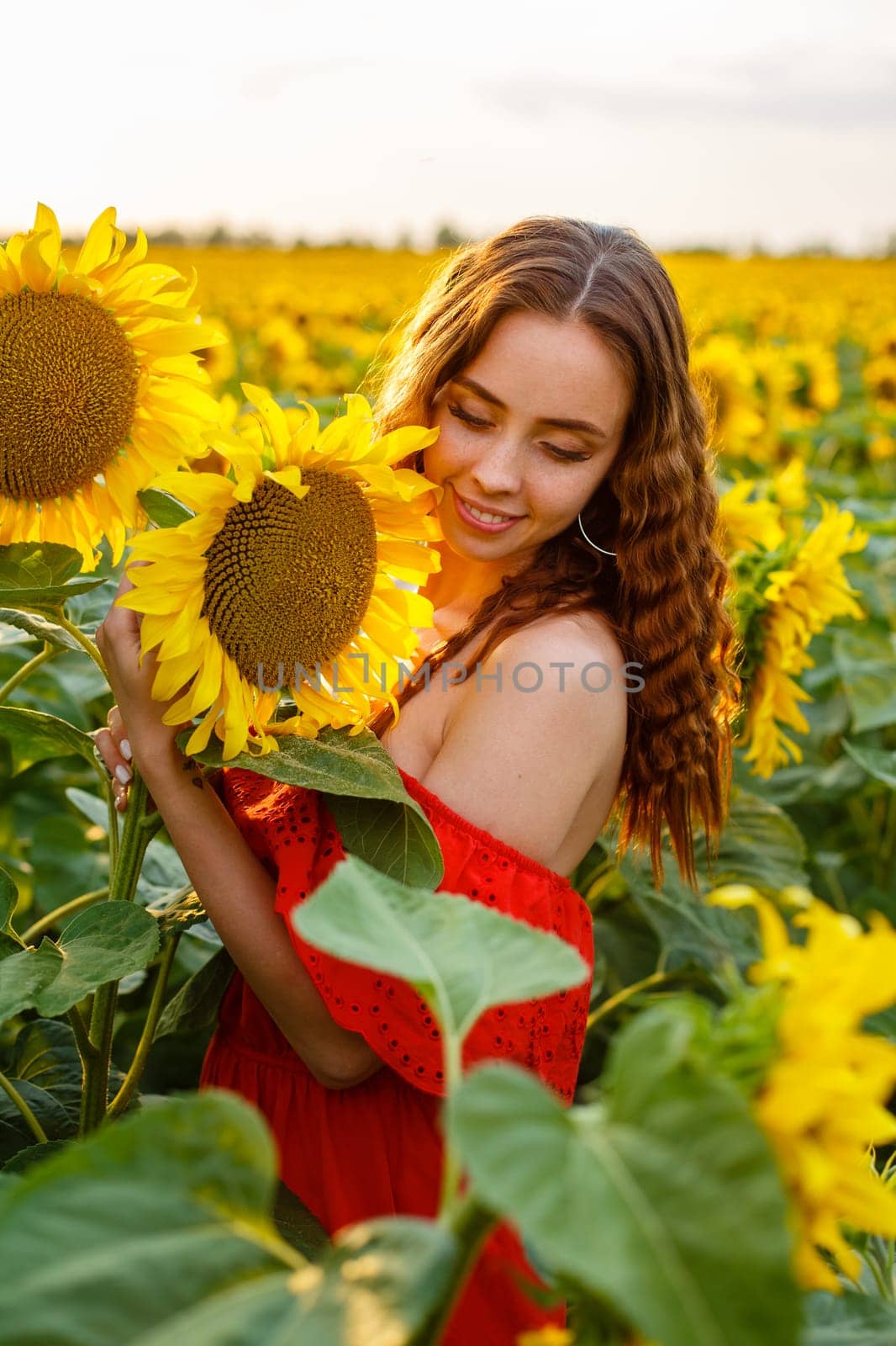 Young woman smiling at sunset in a field of sunflowers. A cute girl of Caucasian appearance in a red dress is happy and free to stand in the evening in the rays. Natural beauty in nature