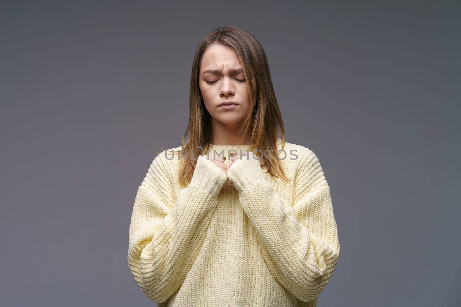 Portrait of a sad young woman of Caucasian ethnicity, clenches her fists in a yellow sweater on a gray background. Experiencing failure and solving problems