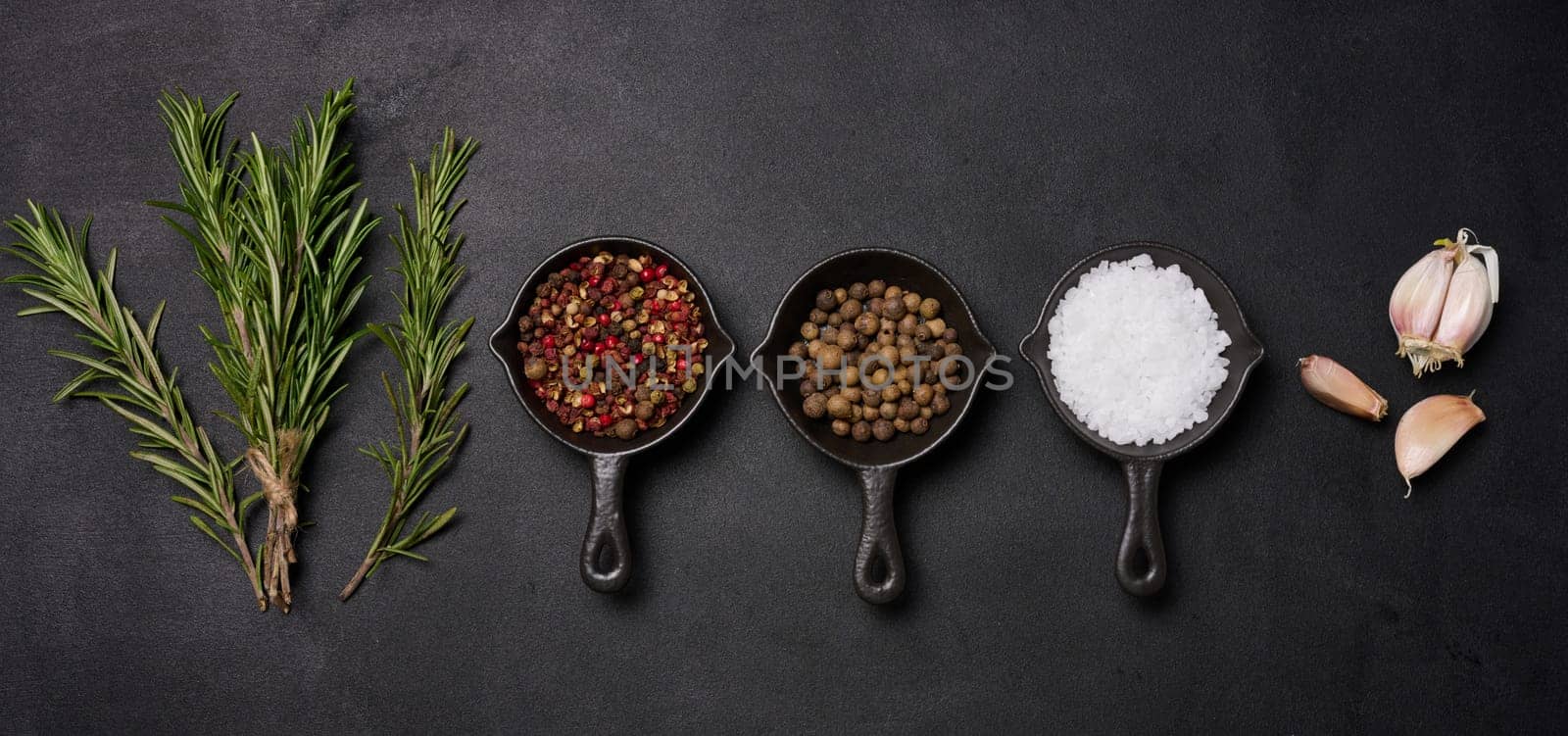 Miniature pans with spices, salt, black pepper and fragrant pepper, a sprig of rosemary on a black table. Spices for cooking by ndanko