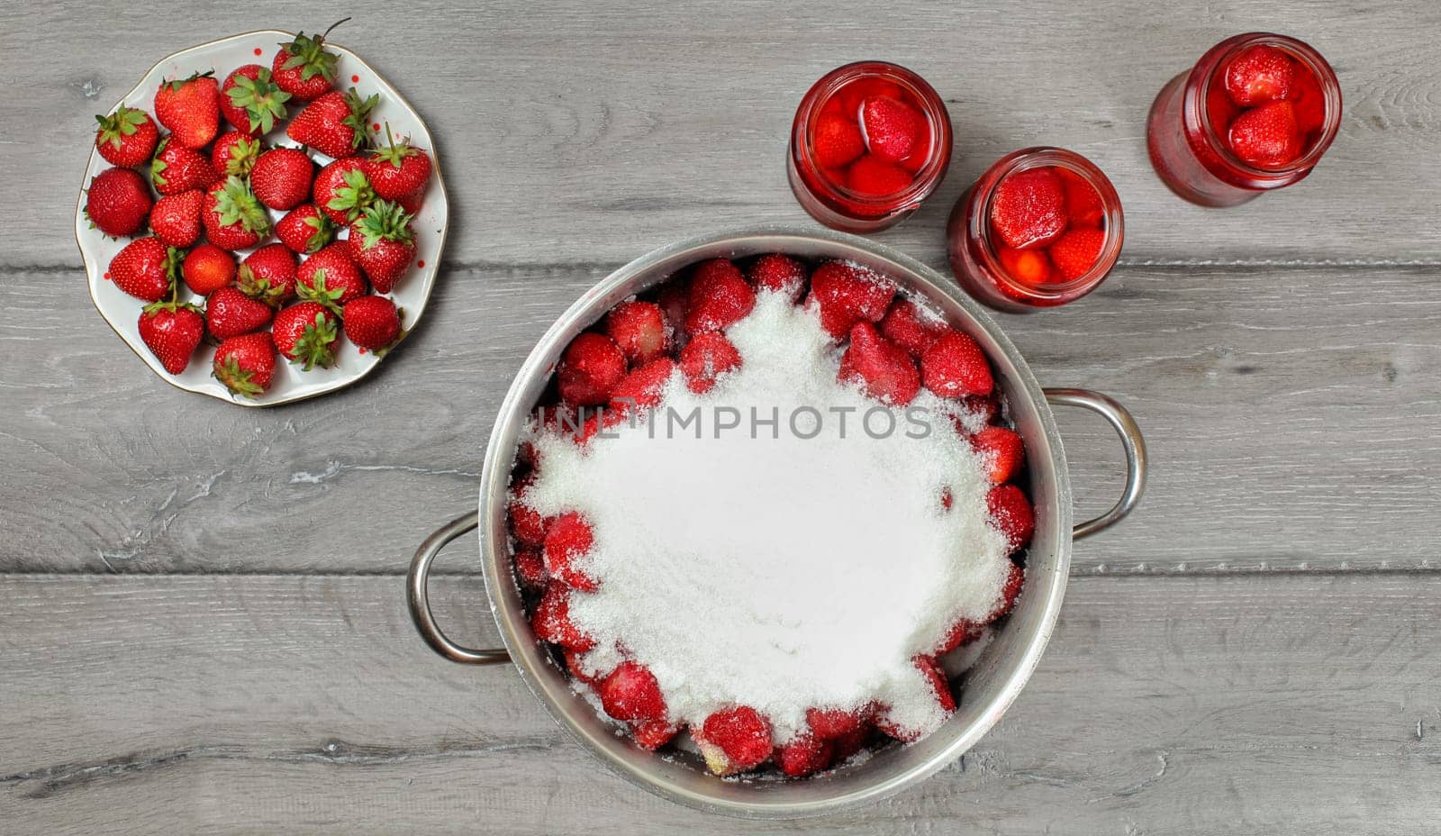 Tabletop photo - large steel pot of strawberries covered with crystal sugar, plate and glass bottles with more fruits around on gray wood desk.