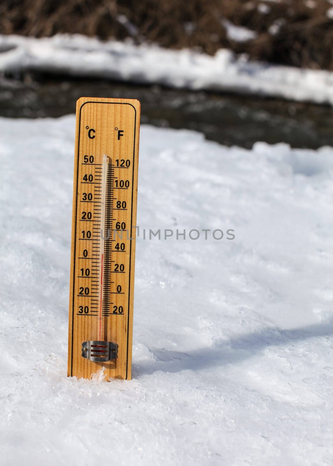 Wooden thermometer standing in ice on partially frozen river, sun shining, showing +3 degrees. Image to illustrate winter leaving, snow slowly thawing, warmer days coming