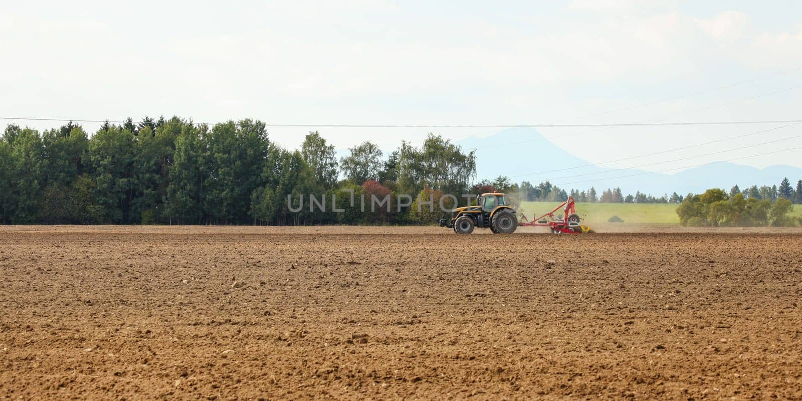 Tractor sowing in empty field on countryside with some mountains in background. by Ivanko