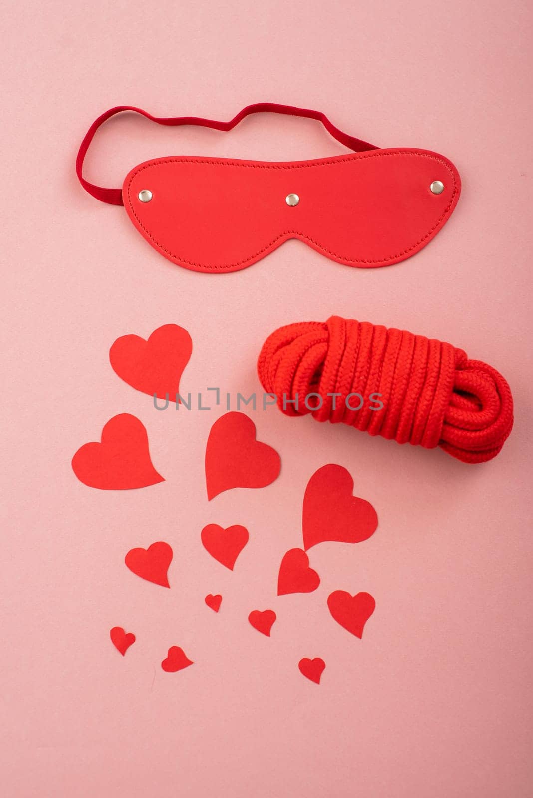 Red leather blindfold mask and rope and hearts on a pink background. Symbol of love and passion