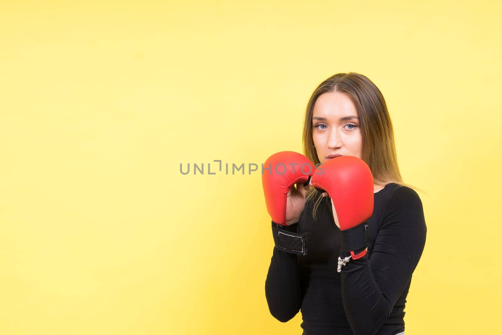 Portrait of young woman in casual clothes and hands in boxing gloves against yellow background by Zelenin