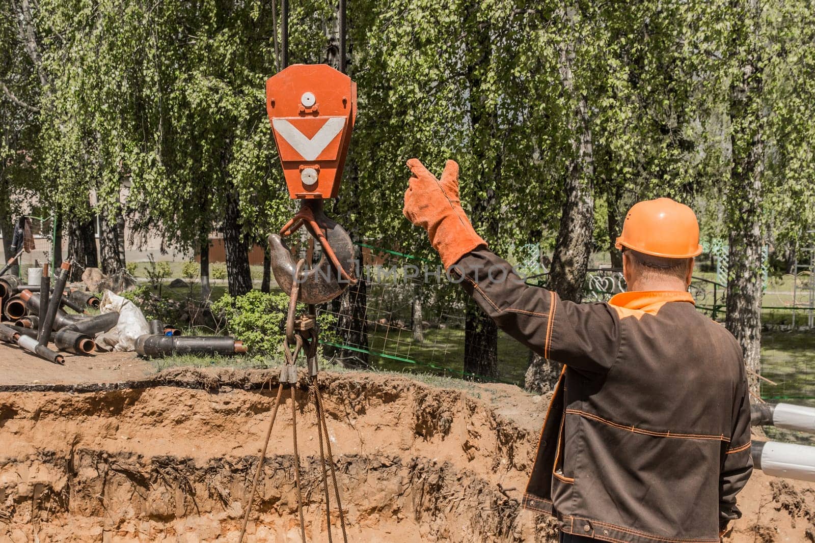 Industrial worker in overalls and protective helmet takes with his hand a heavy hook lifting mechanism of a beam crane on a construction site.