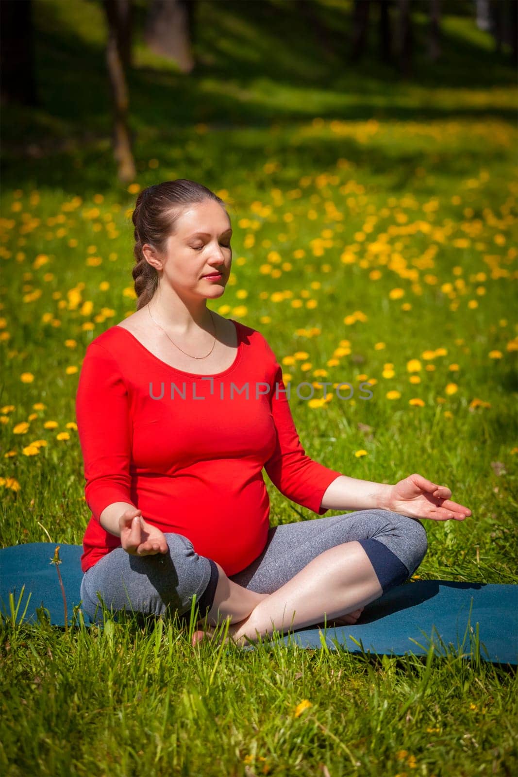 Pregnancy yoga exercise - pregnant woman meditating in yoga asana Sukhasana easy yoga pose with chin mudra outdoors on grass lawn with dandelions in summer