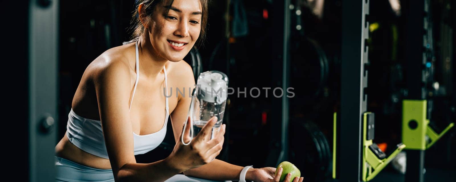 Asian woman holding a green apple and a water bottle in a fitness gym by Sorapop