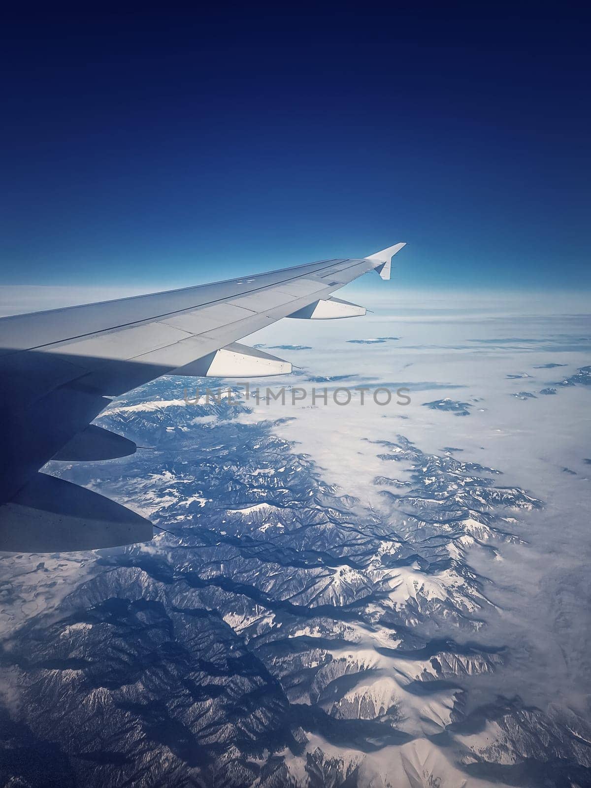 Plane flight above the Carpathian Mountains snowy peaks. Blue skyline and airplane wing seen through the window, vertical background