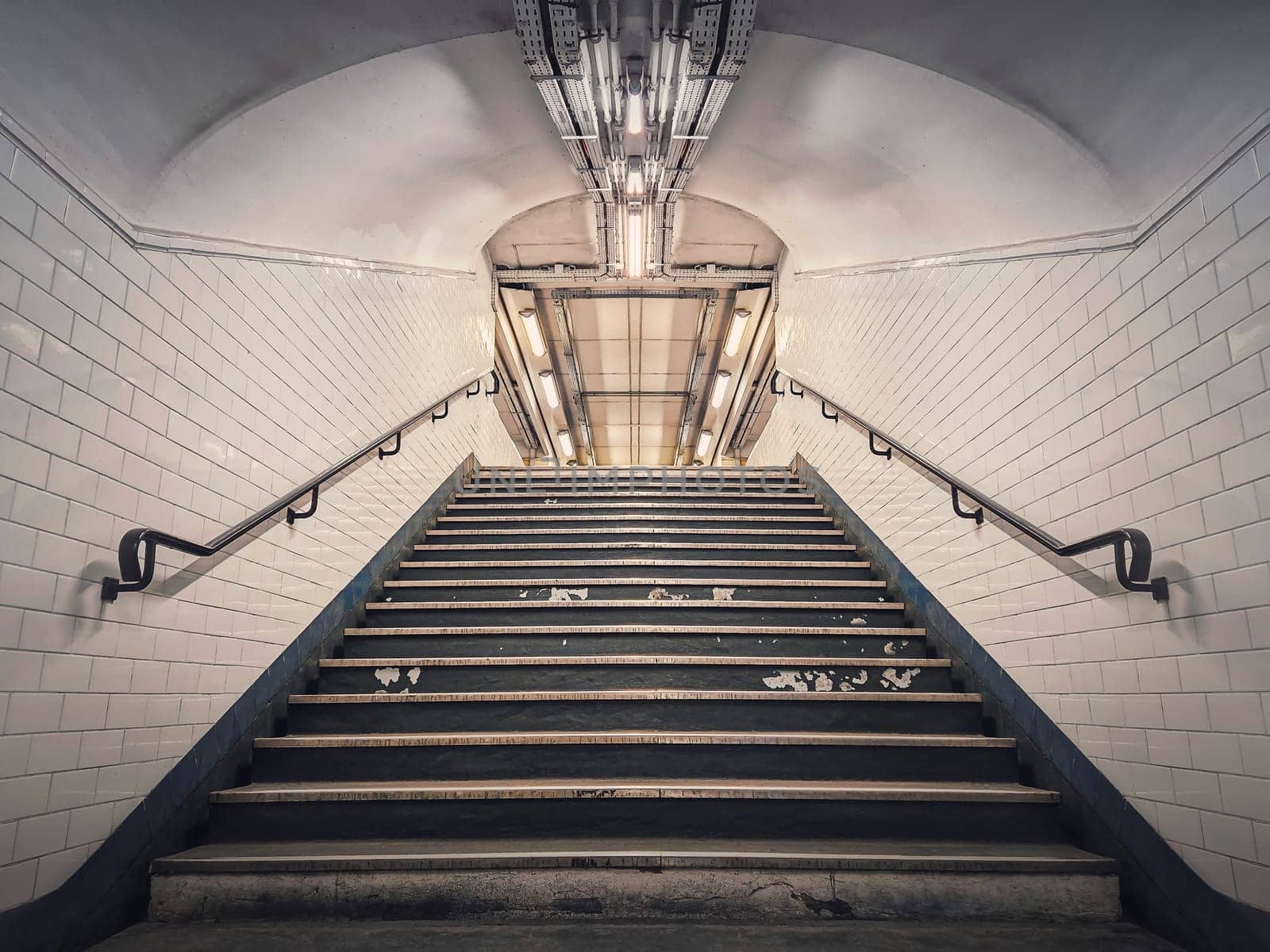 Subway corridor with white tile on walls and glowing lights above a the stairs passage. Symmetrical underground staircase