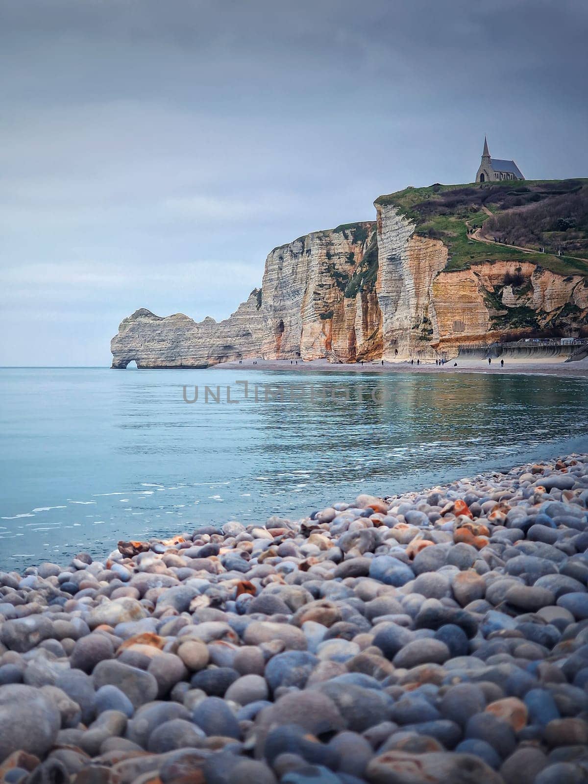 Sightseeing view to Etretat coastline with the famous Notre-Dame de la Garde chapel on the Amont cliff. Pebble beach washed by Atlantic ocean waters, Normandy, France by psychoshadow