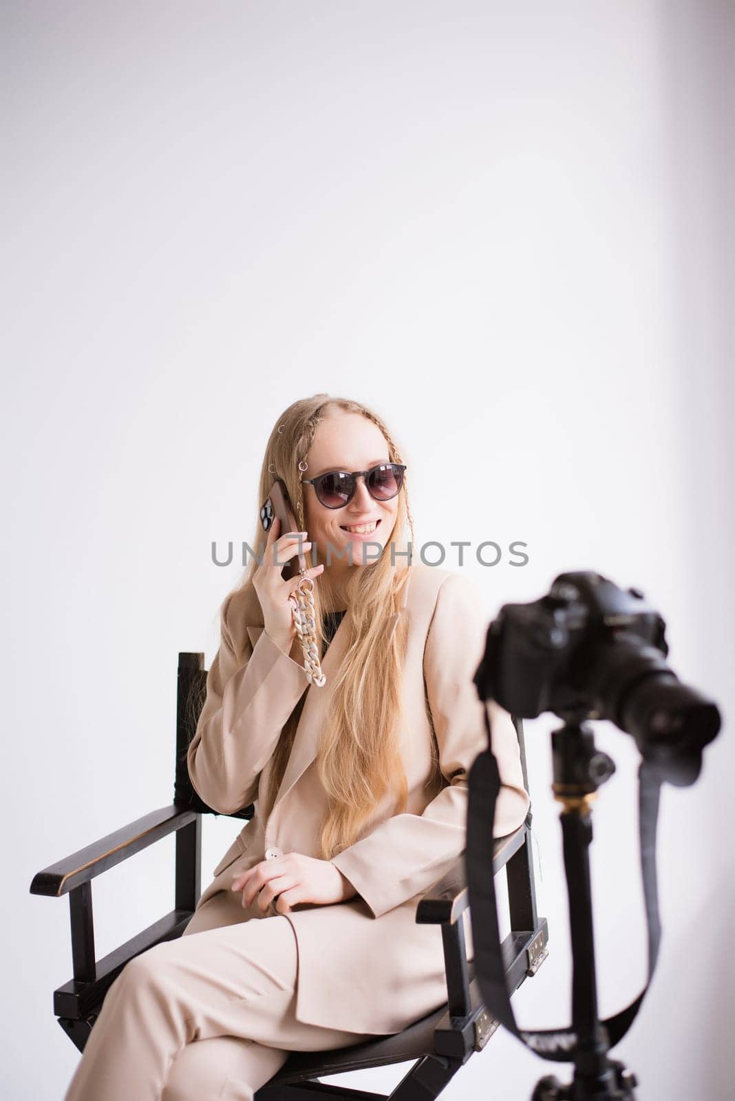 A woman producer videographer, a blonde assistant with a camera and smartphone, happy talking on the phone and smiling in the studio. Wearing a formal nude pantsuit on a white isolated background. Verical