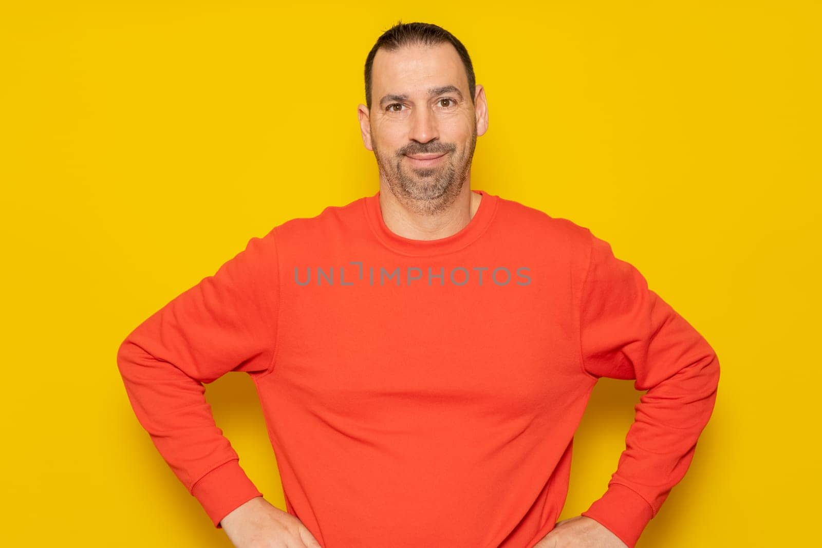 Cheerful bearded man wearing a red sweater posing isolated on gray background, studio portrait. People lifestyle concept. Mock up copy space. Standing with arms akimbo on waist blinking