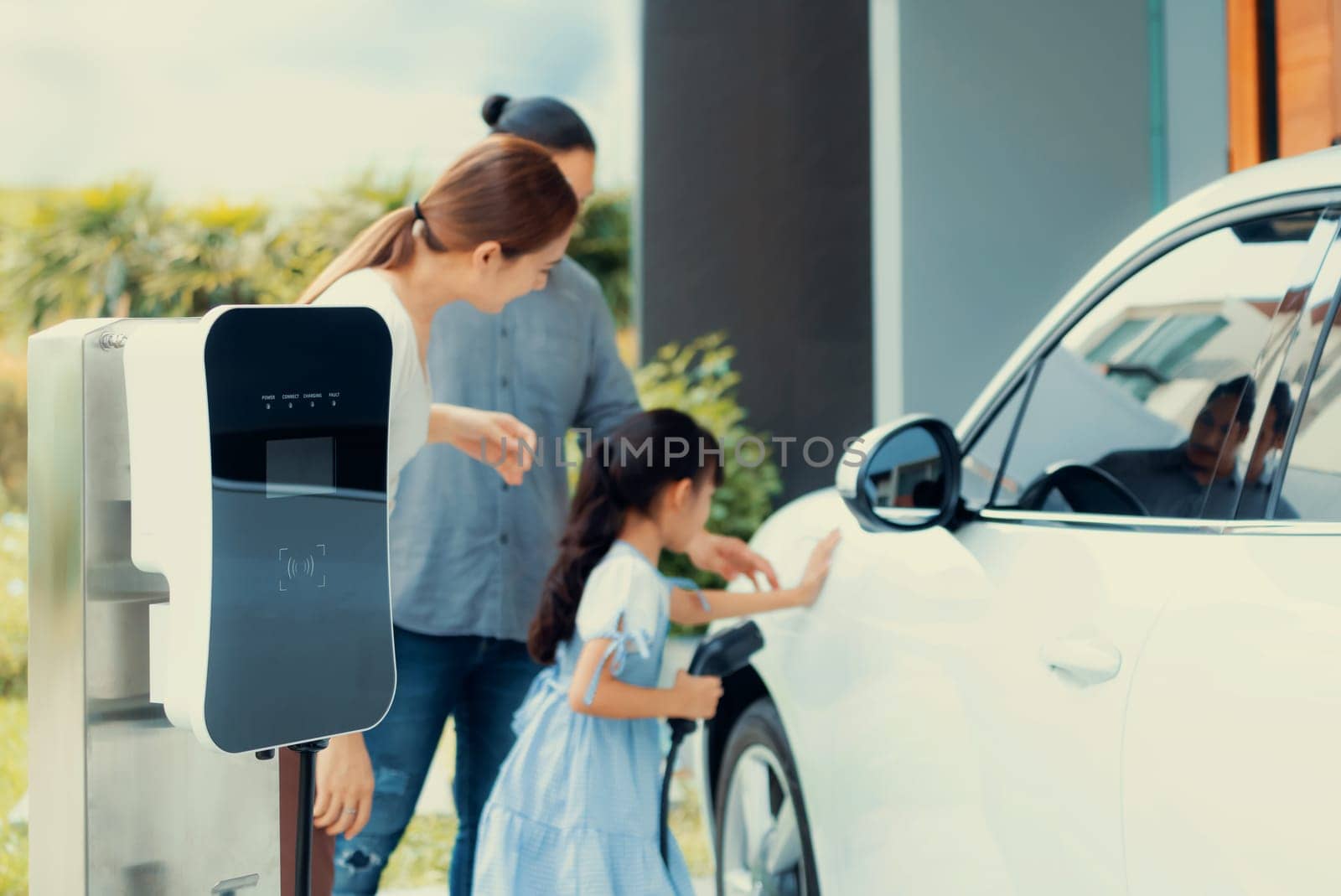 Progressive young parents and daughter with electric vehicle and home charging station. Green and clean energy from electric vehicles for healthy environment. Eco power from renewable source at home.